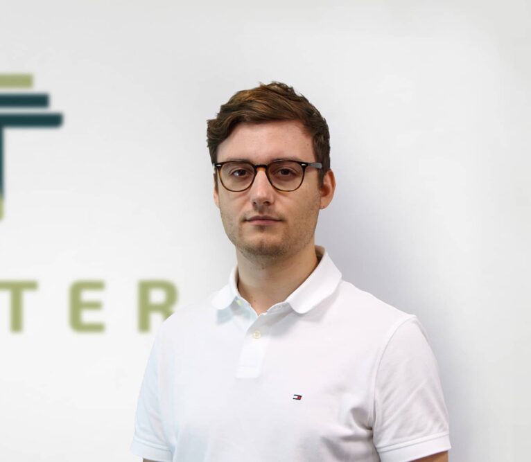 Vīrietis brillēs un baltā polo kreklā stāv priekšā uz balta fona ar daļēji redzamu tekstu un logotipu, smalki norādot uz uzņēmuma zināšanām koka kopnes jomā. Viņam ir īsi brūni mati un neitrāla sejas izteiksme.